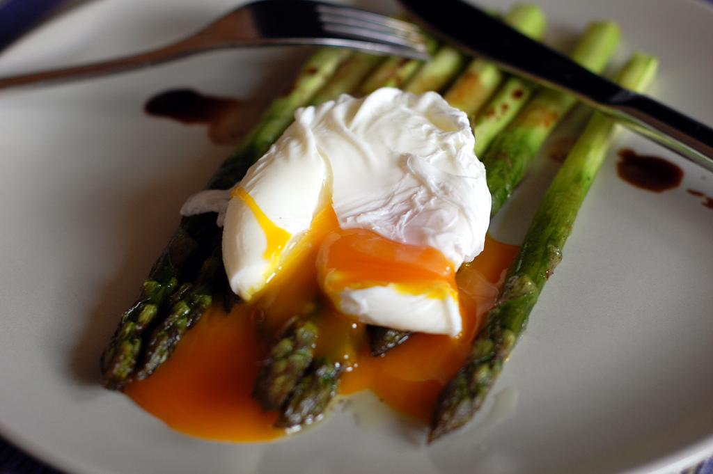 photograph of a runny poached egg atop cooked whole asparagus with a fork and knife in the background.  