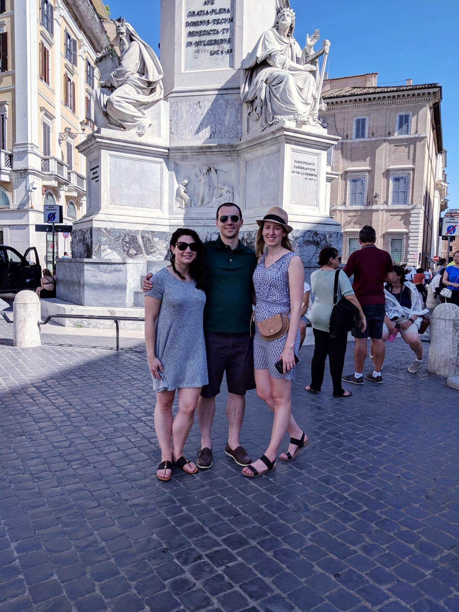Two females and one male posing for a picture in a cobblestone piazza in Rome, Italy. In the background, a white marble obelisk with two statues of males in traditional Roman attire.