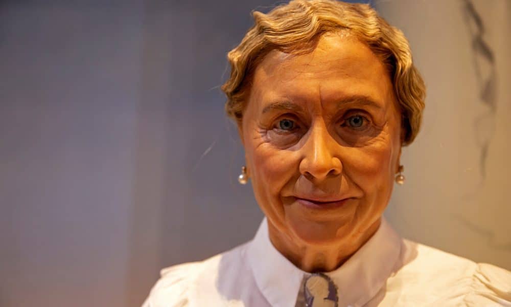 Color photo of Helen Keller as an elderly woman. It is a head shot and her gray hair is pinned back with some waves in the front. She is wearing a white buttoned up shirt and pearl earrings, and she is looking into the camera and smiling