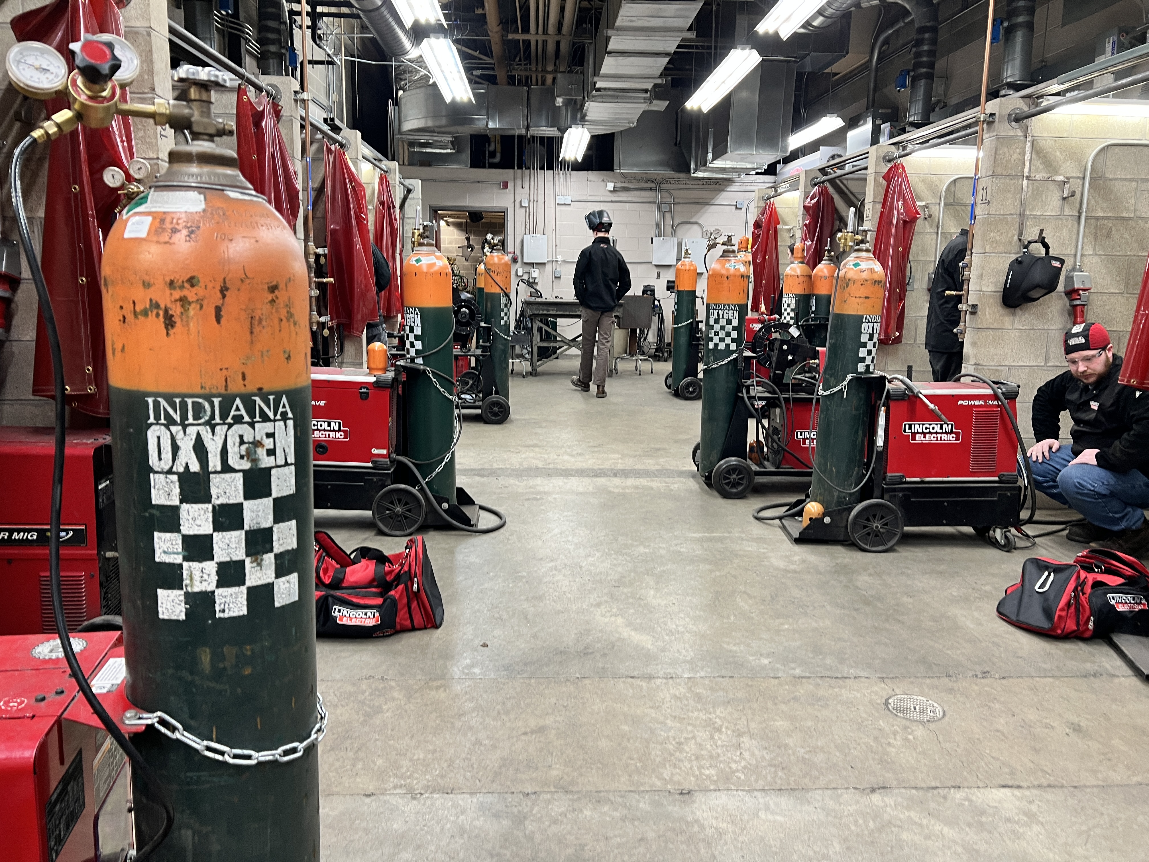 photo of the back of a welding a student walking away the point of view, in a welding lab consisting of six or more gas cylinders, welding machines and booths. 