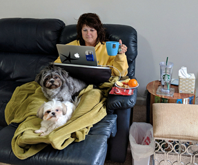 Sandi is sitting on her couch with a blanket over her and two dogs on her legs. She is typing on her computer and has her sick table materials beside her.