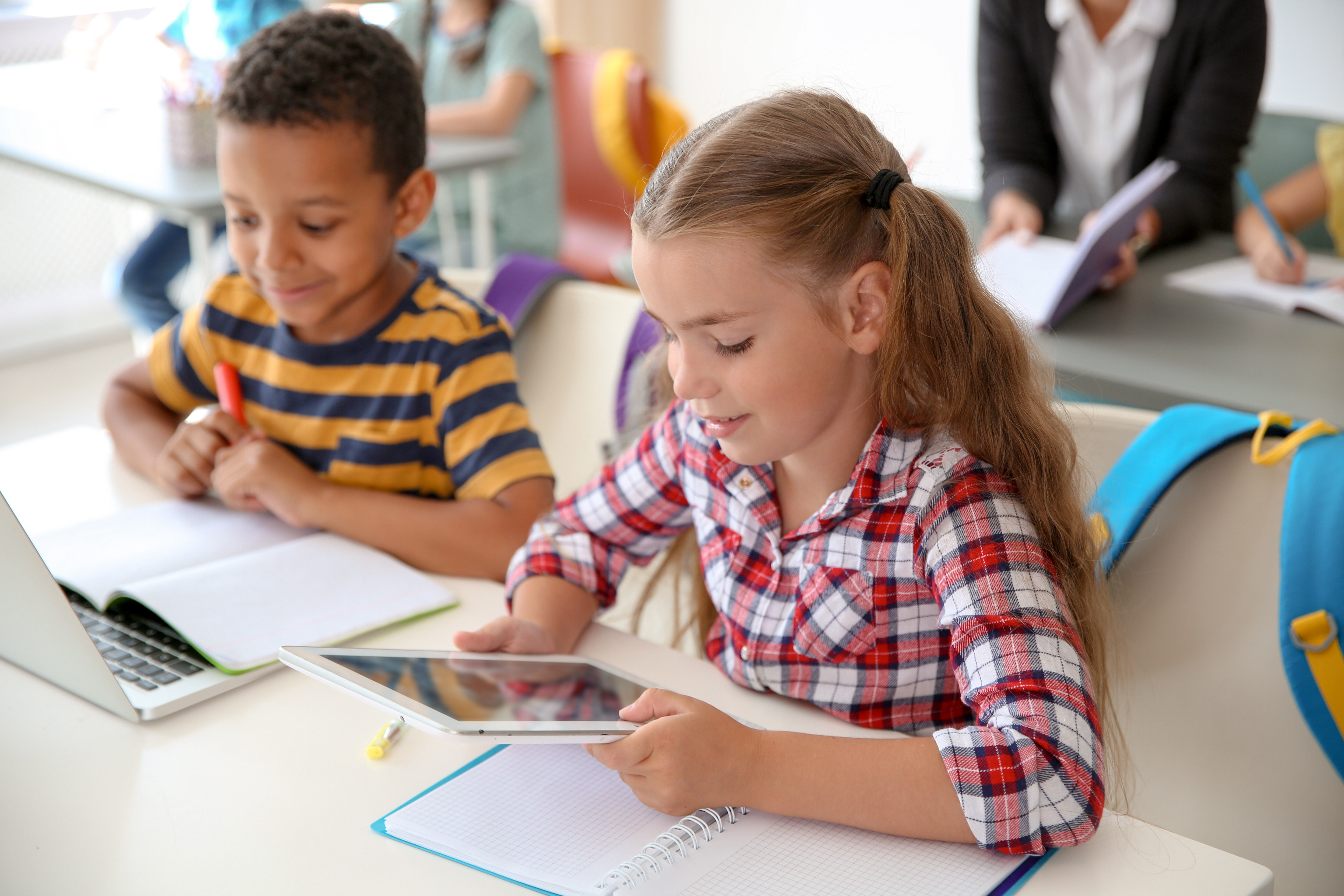 Two second grade students sitting at desk. One is reading on a tablet and the other is reading on a computer and taking handwritten notes.