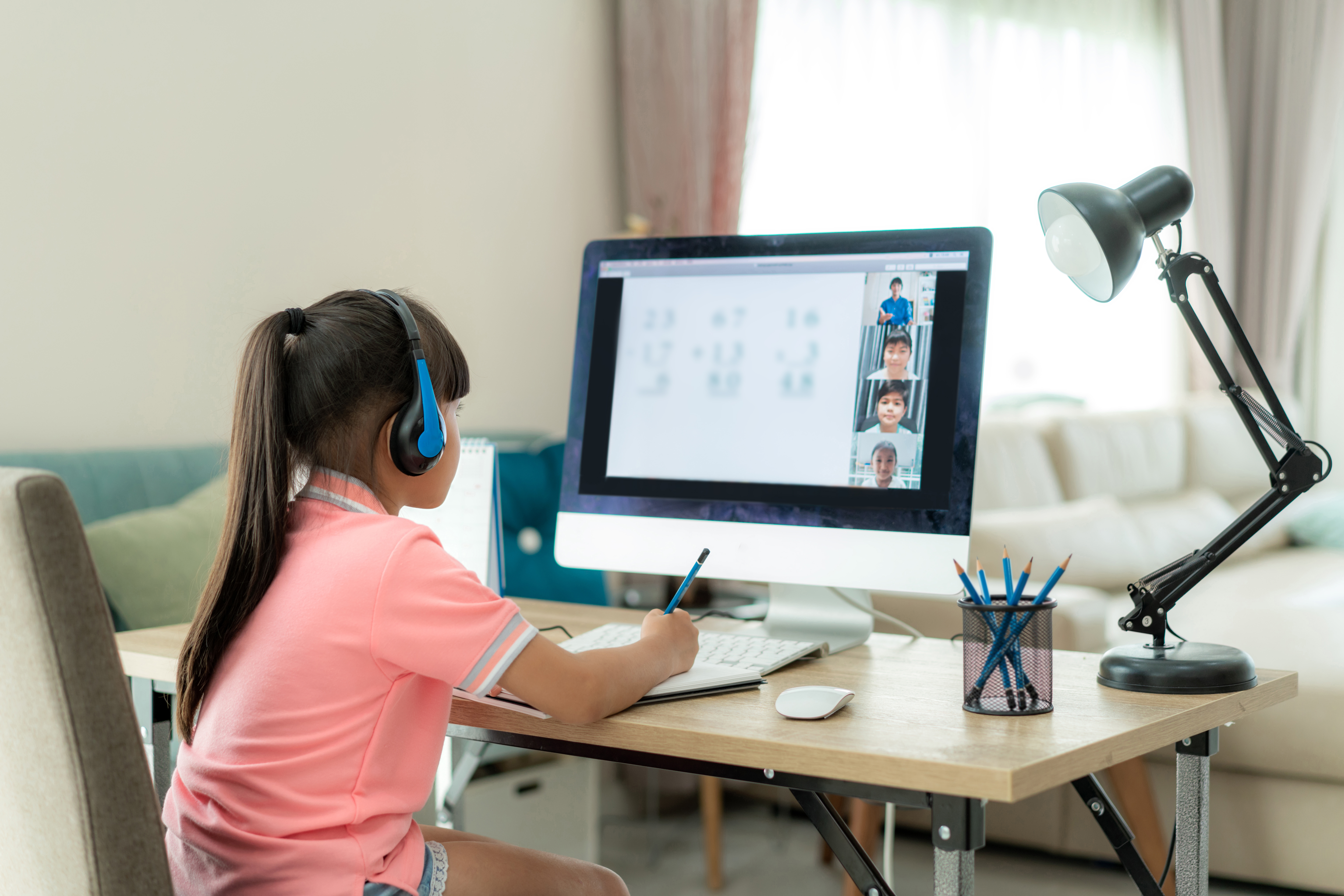 Female student participating in continuous learning from home at at desk. The computer monitor shows class material, teacher and classmates on virtual learning platform.