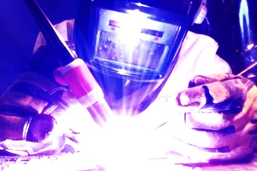close up image of Daniel TIG welding with torch in his right hand and filler metal in his left hand with welding hood and gloves on